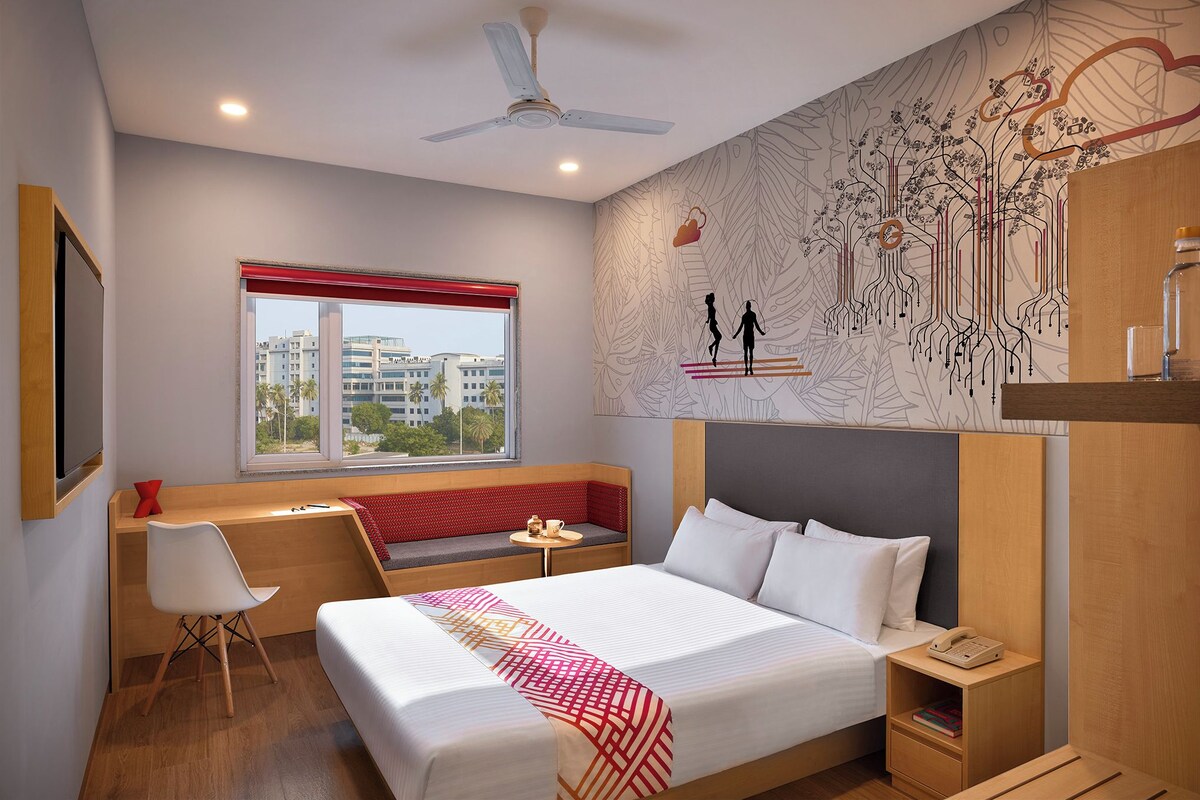 Habitación de hotel con una cama grande, un escritorio con silla, una mesita, un banco, una mesita de noche y una pared con diseño de hojas.