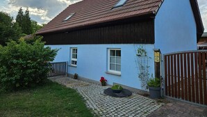 Ein zweistöckiges Haus mit rotem Ziegeldach, blauer Fassade und einem kleinen Gartenbereich mit Topfpflanzen und einem Weg.