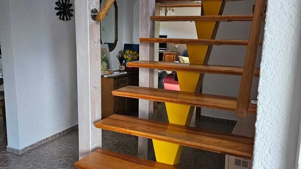 Eine Holztreppe mit abwechselnd gelben und braunen Stufen, die zu einem Podest mit Deckenventilator und Wandverzierungen führt.