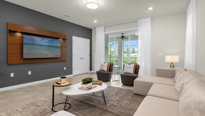 A modern living room with a flat-screen TV mounted on the wall, a beige sofa, a white coffee table with decorative items, and a glass door leading to an outdoor area. - Poolside Oasis · Family Home Close to Disney (Davenport)