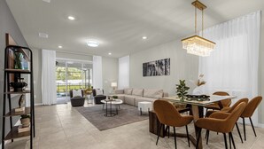 A modern living room with a sofa, coffee table, and dining table with chairs. There is a bookshelf and a chandelier. - Poolside Oasis · Family Home Close to Disney (Davenport)