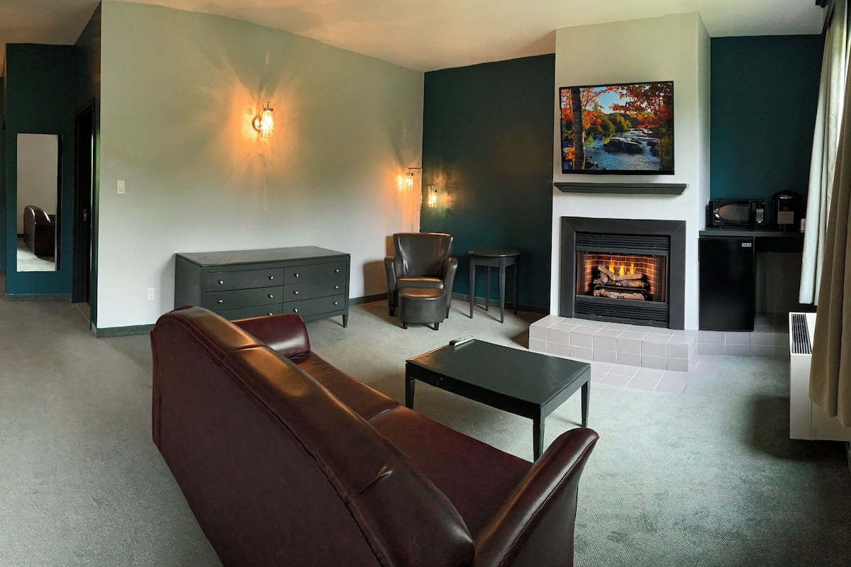 A hotel room with a large bed, a leather sofa, a dining table with chairs, a fireplace, and a view of the dining area.