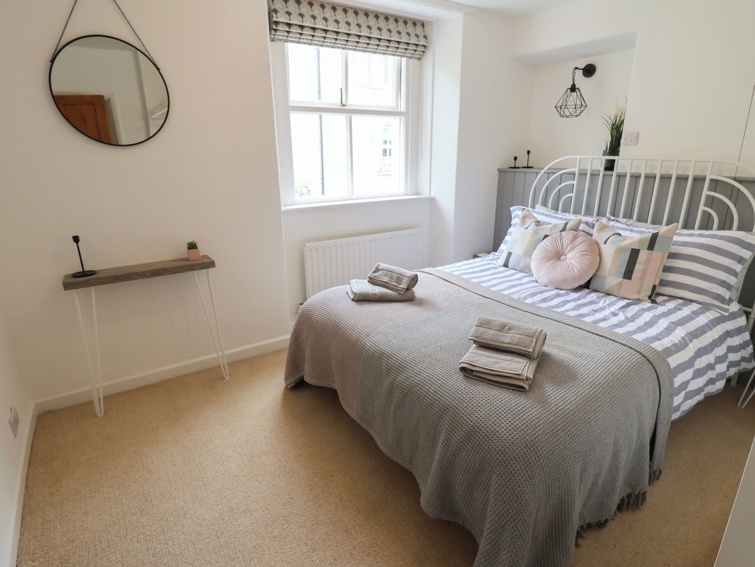 A bedroom with a bed, a bedside table, a hanging mirror, a window with blinds, a radiator, and a wall-mounted light fixture.