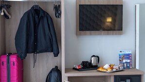 A hotel room with a wooden wardrobe, a desk with a coffee maker, a chair, a pink suitcase, a black backpack, and a television on the wall. - Napoli Forever (Naples)