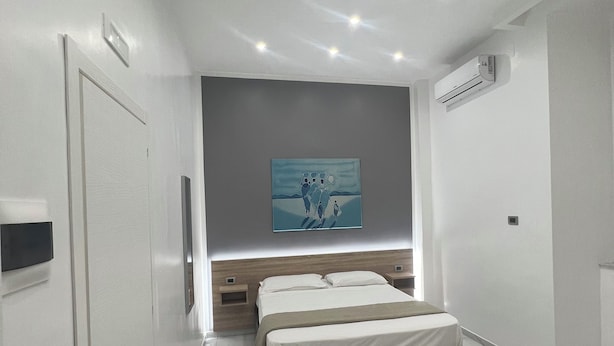 A modern bedroom with a large bed, a headboard with built-in lighting, a painting on the wall, and an air conditioning unit. - Napoli Forever (Naples)