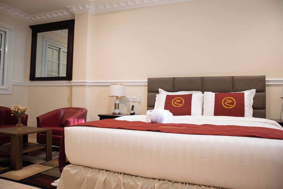 A hotel room with a large bed, red and white pillows, a red throw blanket, a red leather chair, a small table with a vase of flowers, and a nightstand with a lamp.