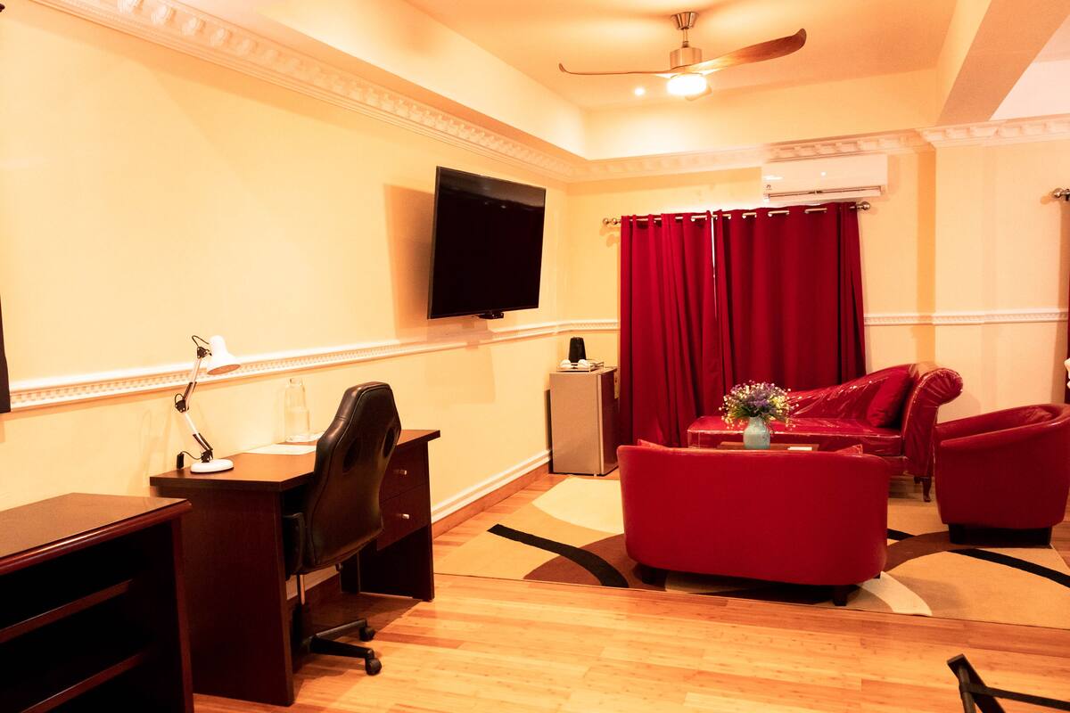A hotel room with a flat-screen TV mounted on the wall, a desk with a chair, a red sofa, and a red armchair.