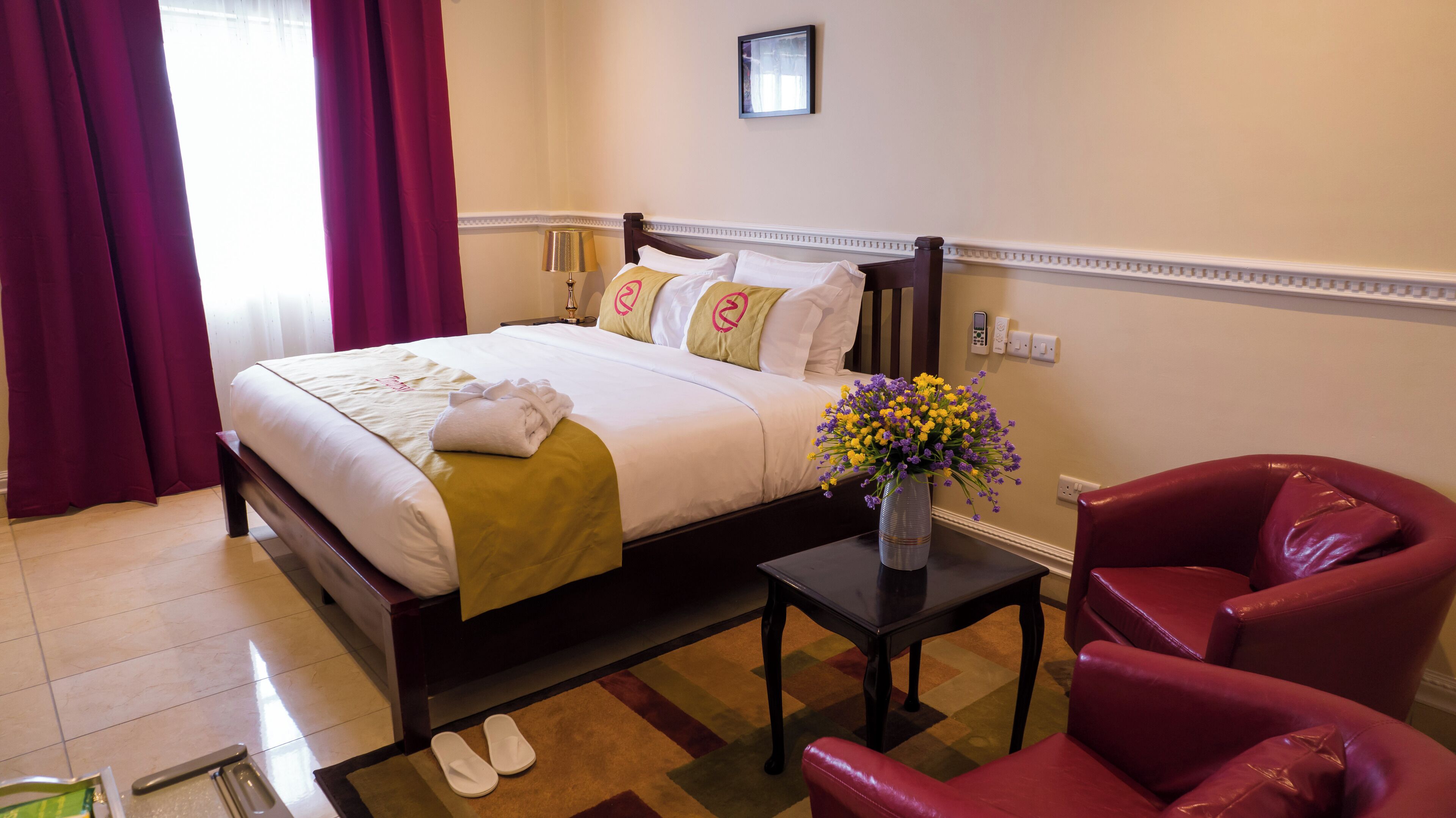 A hotel room with a bed, a small table with a vase of flowers, a red armchair, and a pair of slippers on the floor.