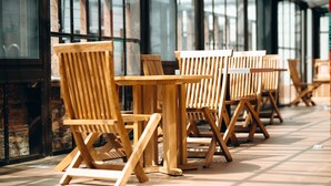 A row of wooden chairs and tables in an outdoor seating area with large windows and a brick wall. - Zimansky Hotel  (Accra)