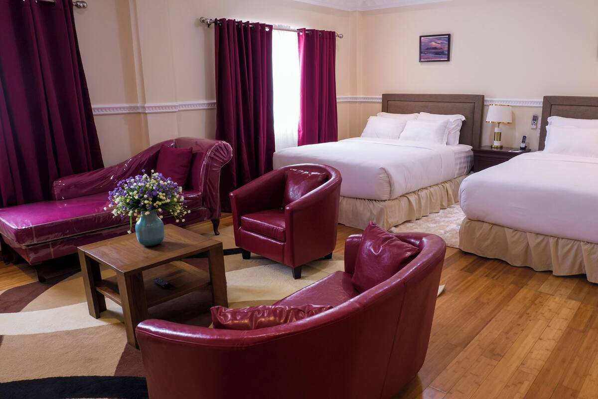A hotel room with two beds, a sofa, a chair, a small table with a vase, and a rug.