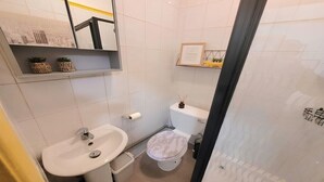 Une salle de bain avec un lavabo, des toilettes et une douche, le tout de couleur blanche. Deux petites plantes en pot sont posées sur une étagère au-dessus du lavabo.