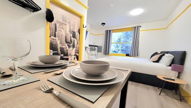 Une chambre d’hôtel avec une table à manger soigneusement dressée, un lit avec une tête de lit noire et une fresque représentant un paysage urbain sur le mur.