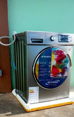 A front-loading washing machine with a front control panel, a water inlet hose, and a label with technical specifications. - Sol y Mar, Galápagos (San Cristóbal)