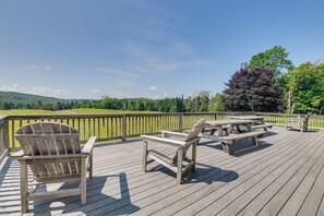 Una terraza de madera con sillas Adirondack y mesas de picnic, con vistas a un paisaje verde y colinas.