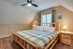 Un dormitorio con una cama de madera, una mesita de noche, un ventilador de techo, una ventana con cortinas y un cuadro en la pared.