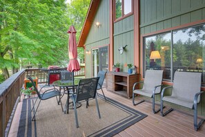 Un patio techado con mobiliario exterior, una mesa y sillas, rodeado por una terraza de madera y ventanas amplias.