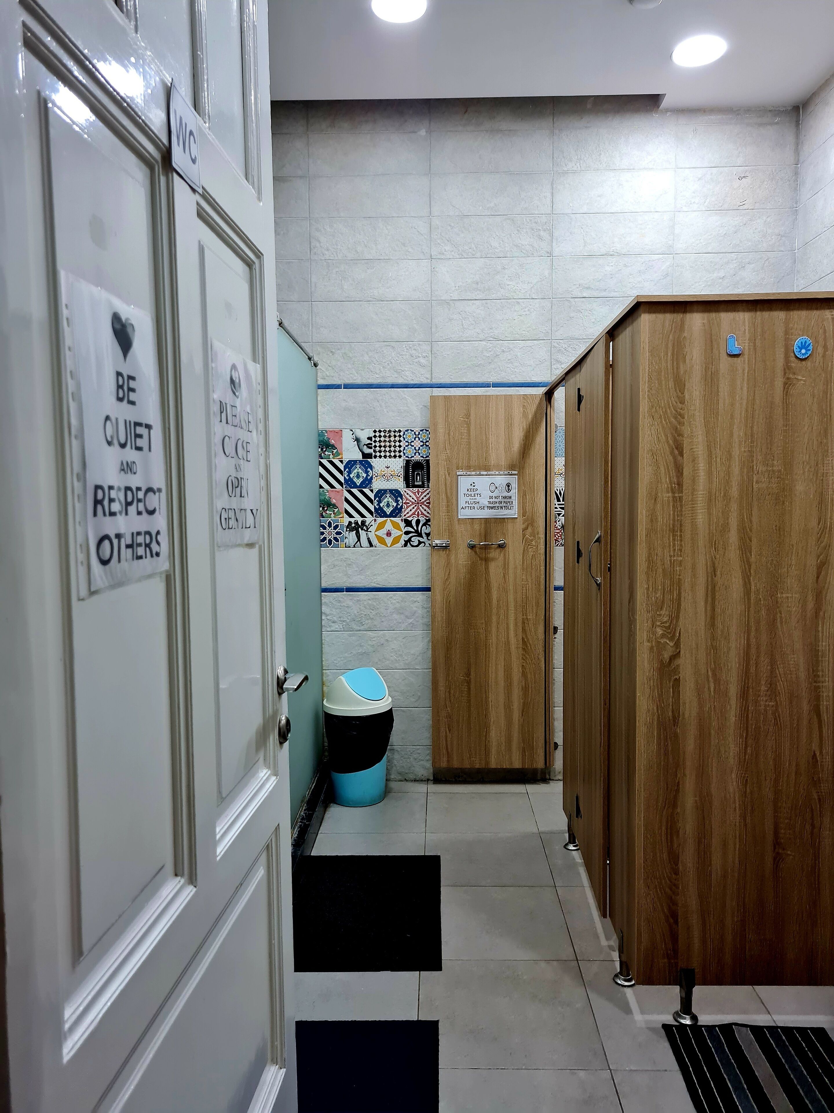 A hallway with WC signs, a sign requesting quiet and respect, a tiled wall, a wooden door, and a trash bin.