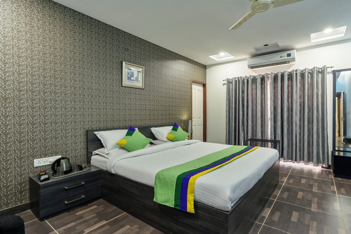 A hotel room with a patterned wallpaper, a bed with a white and green bedspread, a dark wooden nightstand, a black drawer, a black chair, a ceiling fan, and an air conditioning unit.