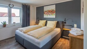 Een tweepersoonsbed met een grijs hoofdeinde, een nachtkastje met een lamp en een raam met gordijnen.