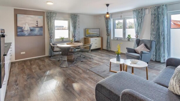 Een moderne woonkamer met een grijze bank, een eettafel met stoelen en een kleine salontafel. Er hangt een schilderij aan de muur, er is een televisie en een raam met gordijnen.
