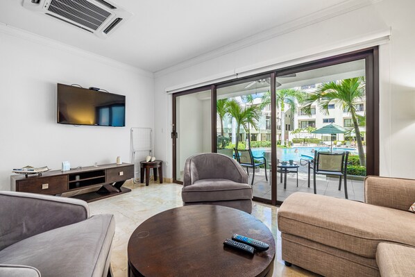 A modern living room with a flat-screen TV, a sofa, a coffee table, and a sliding glass door leading to a pool area.