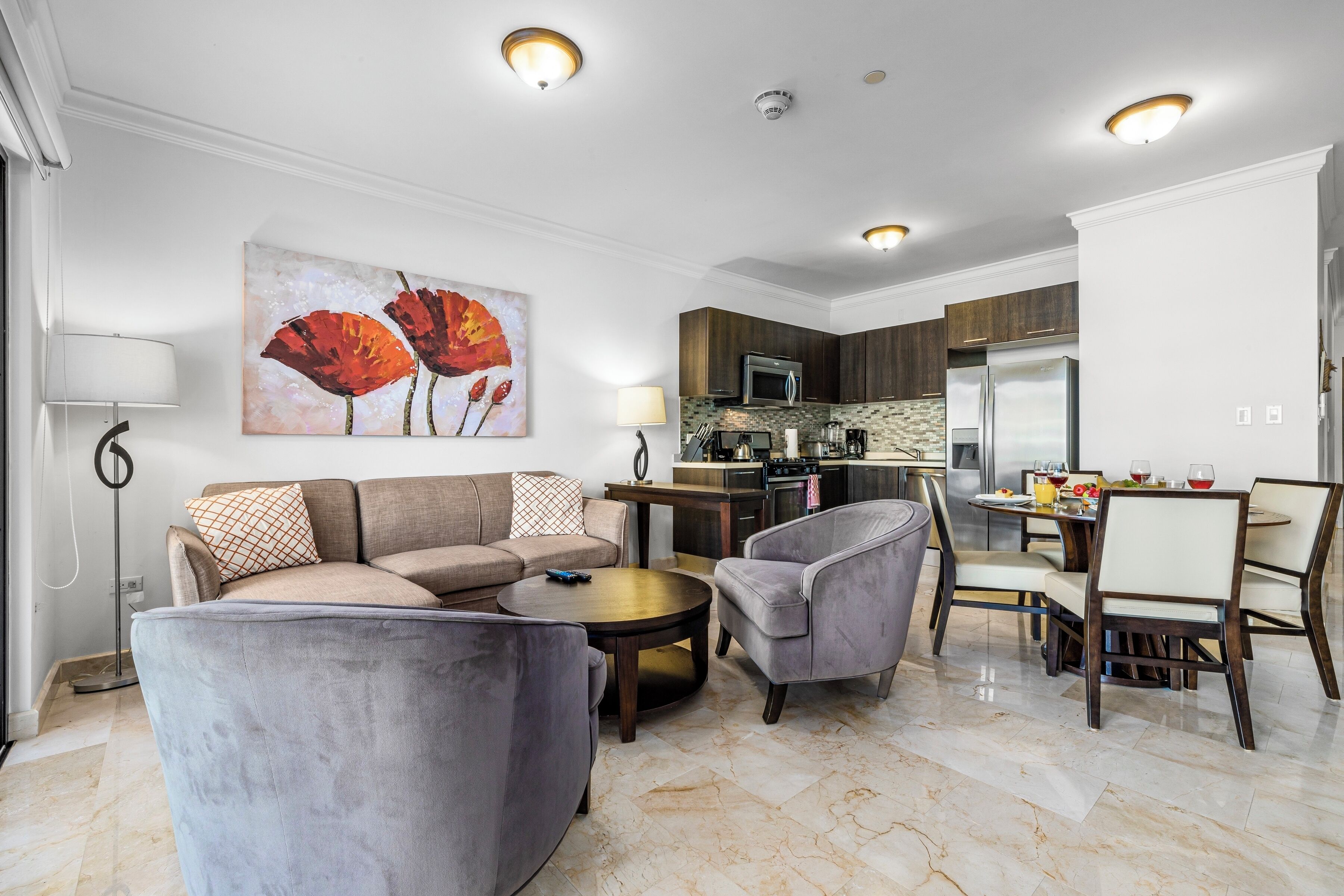 Una sala de estar moderna con un sofá, sillones y una mesa de centro. Se observa un comedor con una mesa y sillas. La cocina se ve al fondo, con electrodomésticos y armarios.