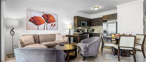 Una sala de estar moderna con un sofá, sillones y una mesa de centro. Se observa un comedor con una mesa y sillas. La cocina se ve al fondo, con electrodomésticos y armarios.