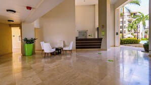 A hotel lobby with a reception desk, a seating area with chairs, a large green planter, and a view of a residential building with balconies. - Condo 1 one bed, Palm Beach (Noord)