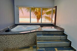 Un baño con una bañera grande, un muro con mosaicos de azulejos y vista a la playa a través de un ventanal amplio.