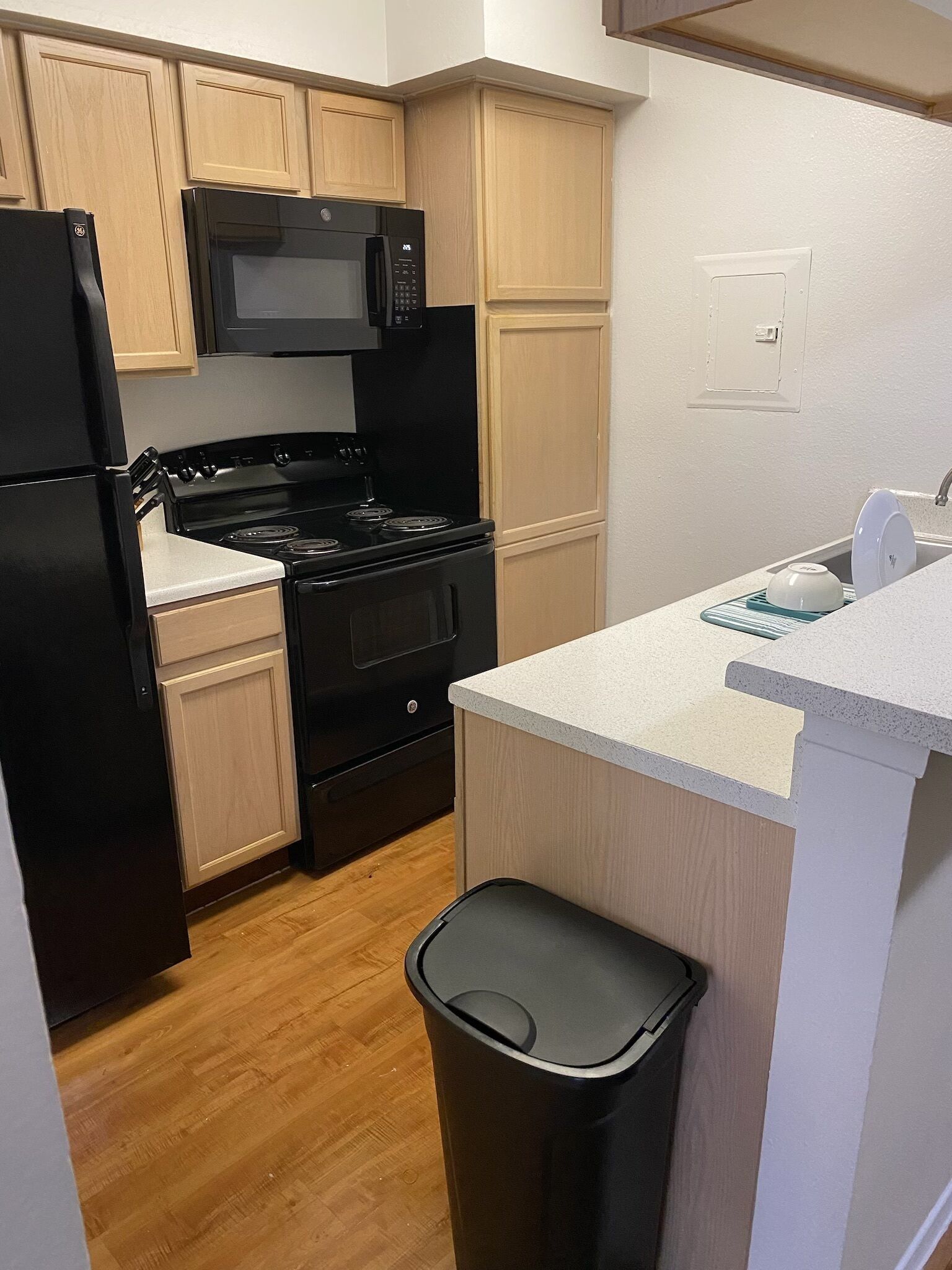 A compact kitchen with black appliances, wooden cabinets, and a small trash bin.