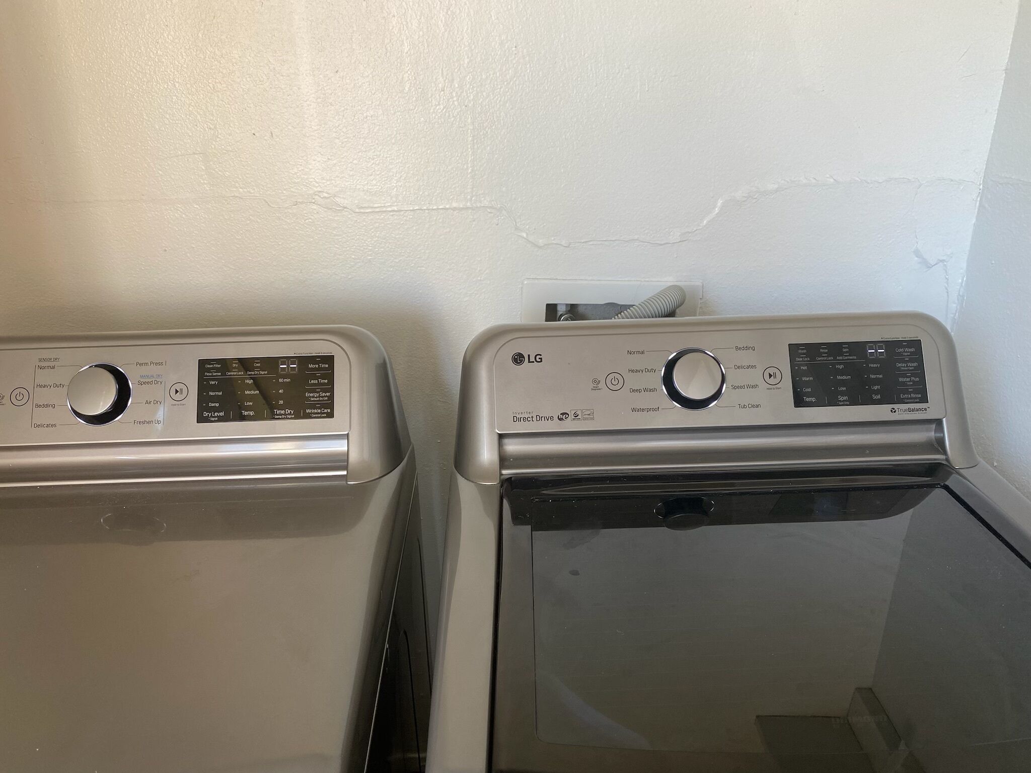Two front-loading washing machines in a laundry room with a white wall and a visible crack.