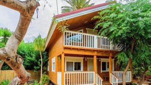 Une maison en bois à deux étages, dotée d’un balcon, entourée d’une végétation luxuriante et agrémentée de plantes en pot.