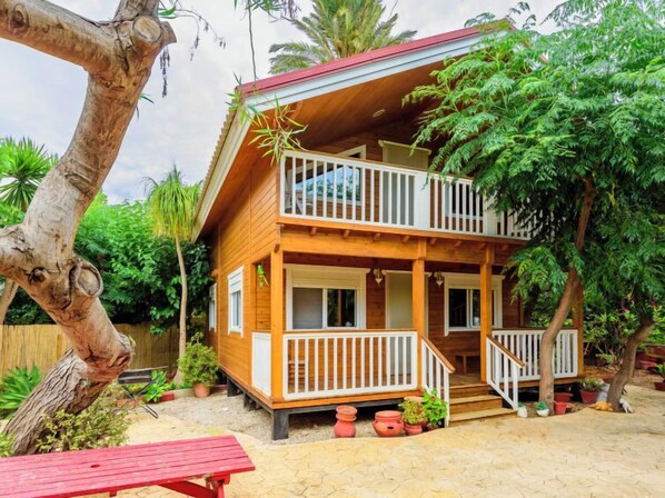 A two-story wooden house with a balcony, surrounded by lush greenery and potted plants.