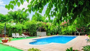Une piscine rectangulaire, bordée d’une terrasse carrelée et entourée d’une végétation luxuriante et de plantes en pot.