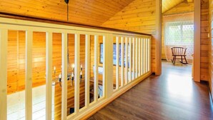 Un intérieur en bois avec un plafond haut, une balustrade blanche et une vue sur un coin repas où se trouve un fauteuil à bascule.