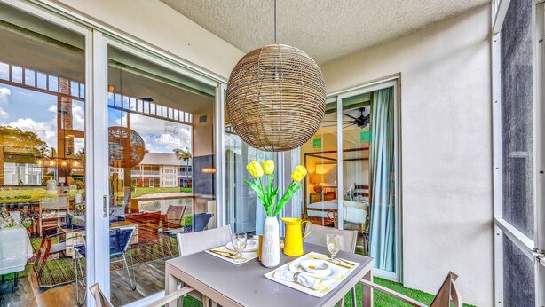 Un área de comedor al aire libre con una mesa lista para dos personas, una lámpara colgante de mimbre y un jarrón con flores amarillas.
