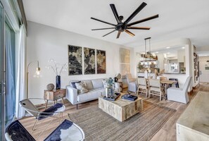 A modern living room with a sofa, coffee table, and chairs. There is a dining area with a table and chairs. The room has a ceiling fan and artwork on the wall.