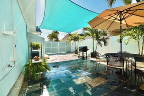 Eine Terrassenfläche mit gefliestem Boden, Gartenmöbeln, einem Tisch und Stühlen sowie einem großen Sonnenschirm.