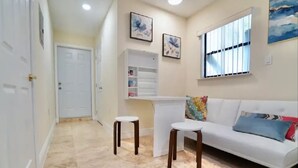 Ein Wohnzimmer mit einem weißen Sofa, zwei weißen Hocker, einem bunten Kissen, einem weißen Schrank und einem Fenster mit Jalousien.