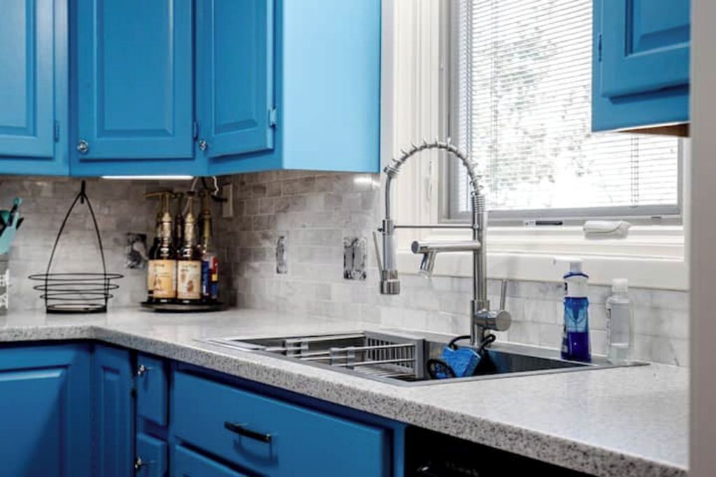 Une cuisine avec des meubles de couleur bleue, un évier en acier inoxydable et une fenêtre avec des stores.