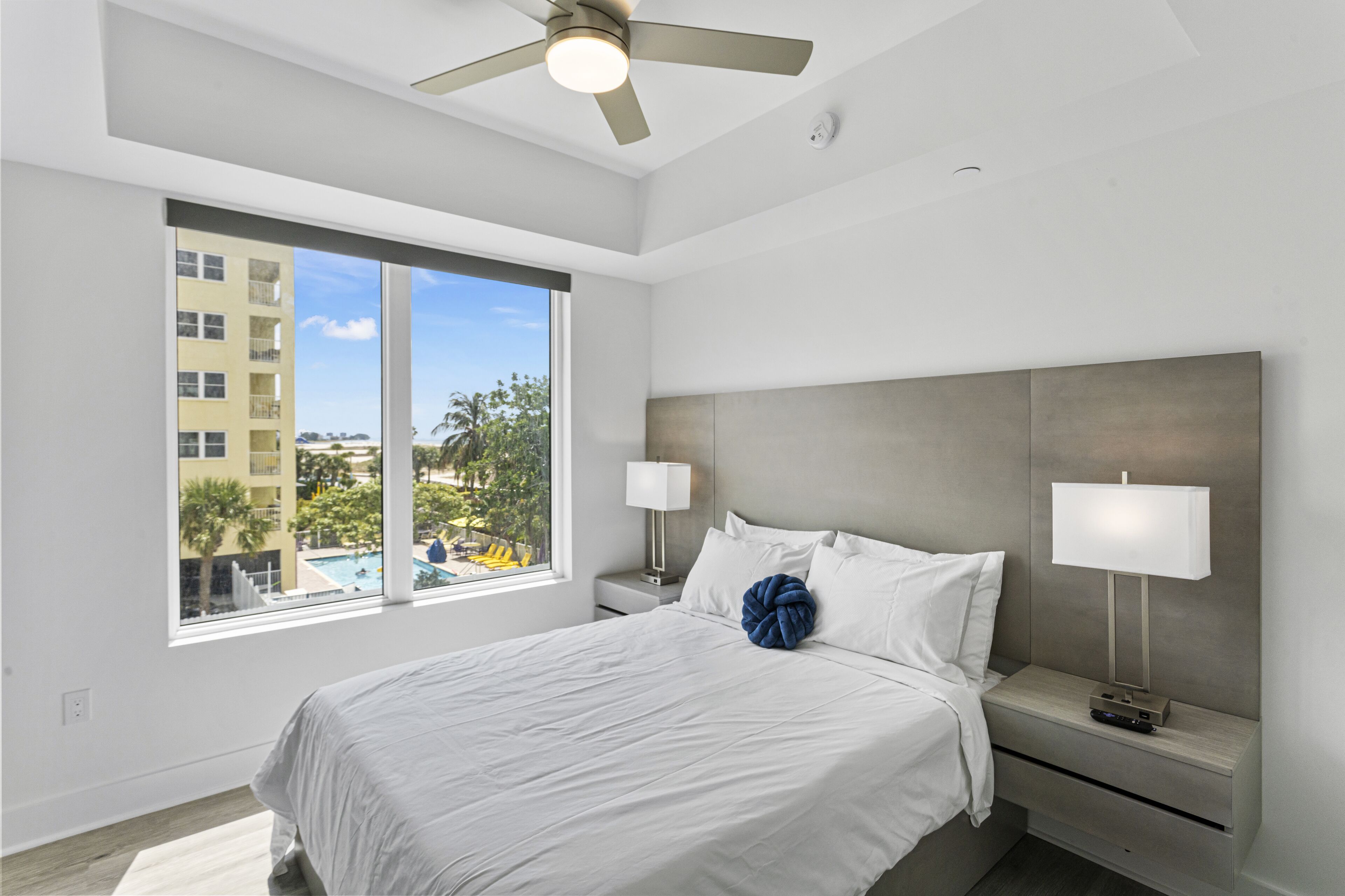 A modern hotel room with a large bed, two bedside tables, a ceiling fan, and a window with a view of buildings and trees.
