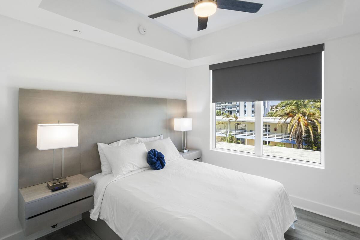 A modern hotel room with a large bed, two bedside tables, a ceiling fan, and a window with a view of buildings and palm trees.