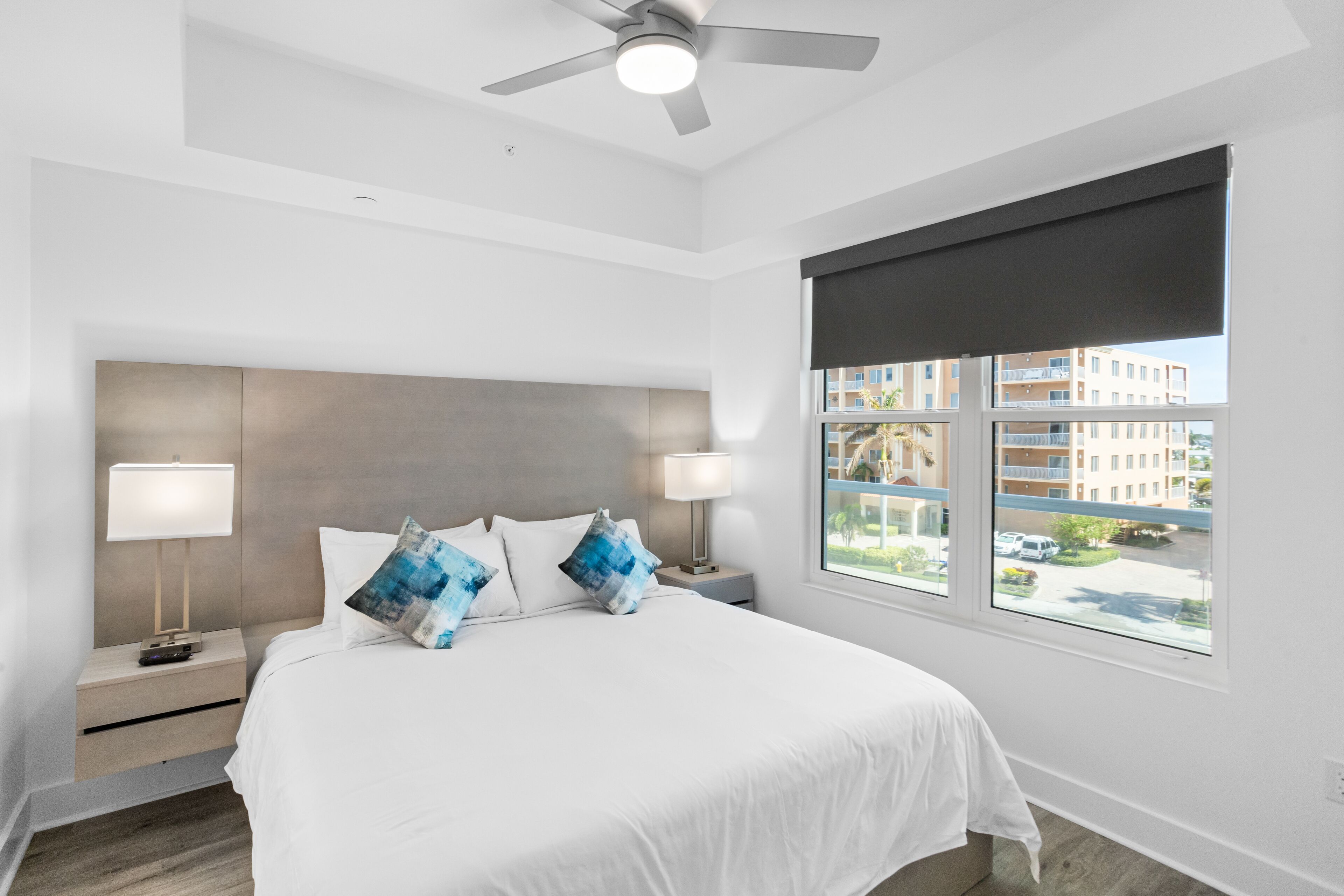 Habitación de hotel moderna con una cama grande, dos mesitas de noche con lámparas, un ventilador de techo y un ventanal con vistas a edificios.