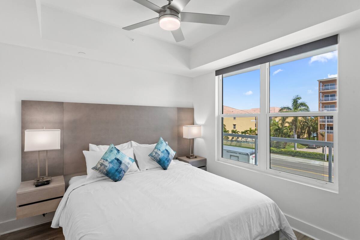 A modern hotel room with a large bed, two bedside tables with lamps, a ceiling fan, and a large window offering a view of the city.