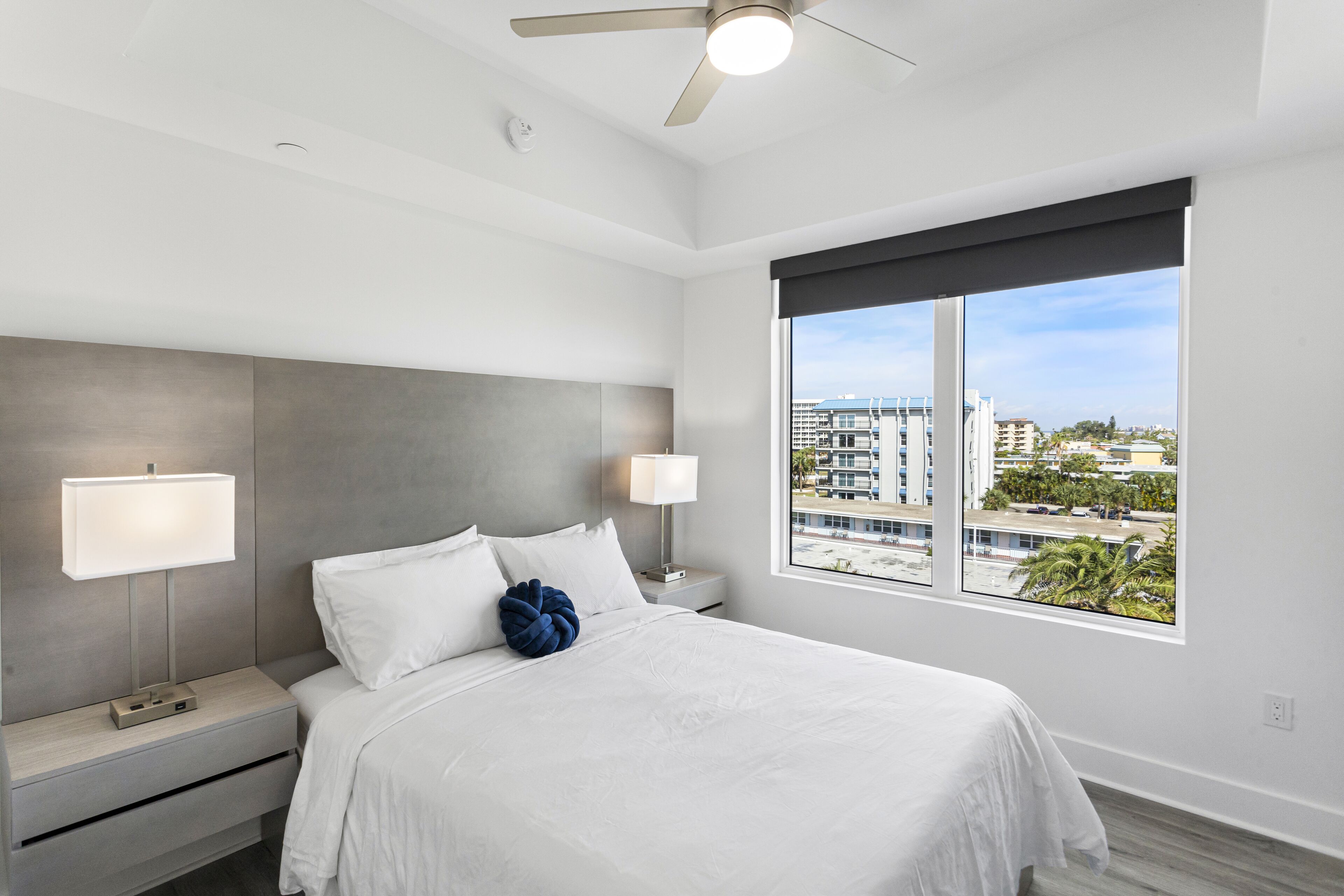 Une chambre d’hôtel moderne dotée d’un grand lit, de deux tables de chevet avec lampes, d’un ventilateur de plafond et d’une grande fenêtre donnant sur la ville.
