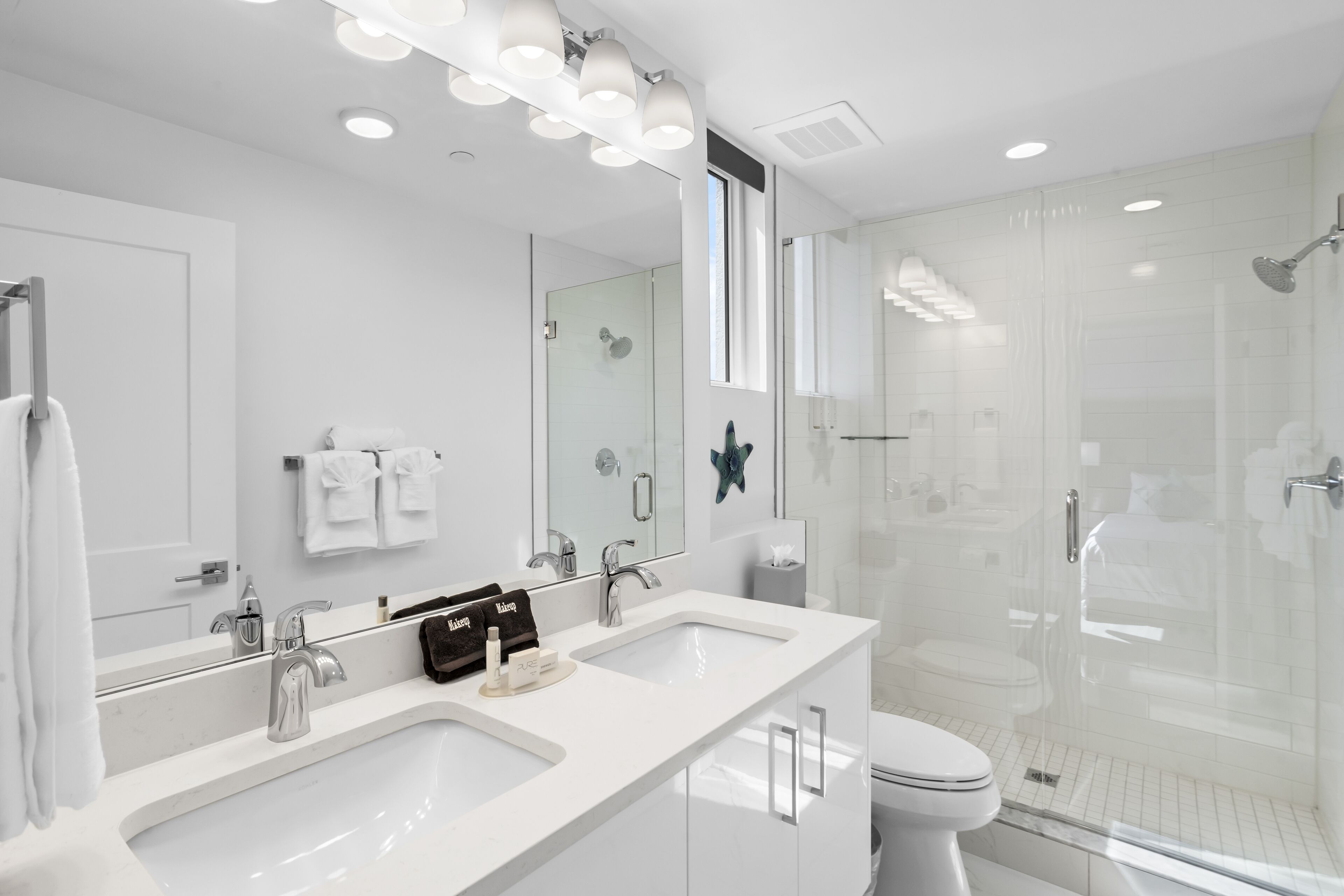 A modern bathroom with a large mirror, two sinks, a toilet, and a glass-enclosed shower.