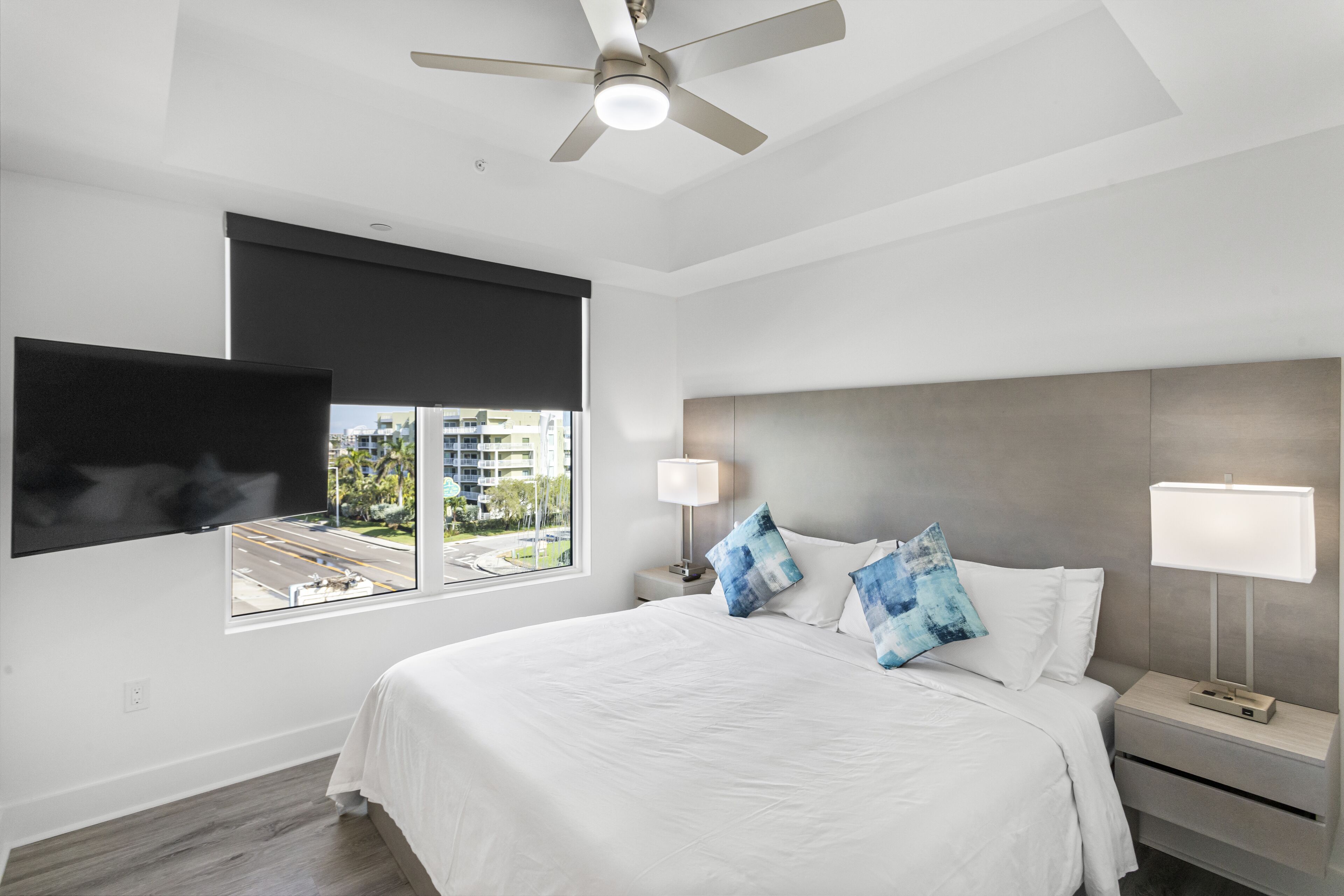 Una habitación de hotel moderna con una cama grande, un ventilador de techo, un televisor montado en la pared, una ventana con vistas a edificios y una mesita de noche con lámpara.