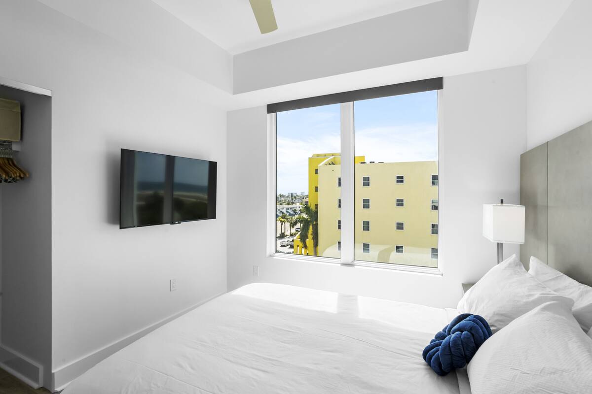 A modern hotel room with a large bed, a flat-screen TV mounted on the wall, and a window offering a view of a yellow building.