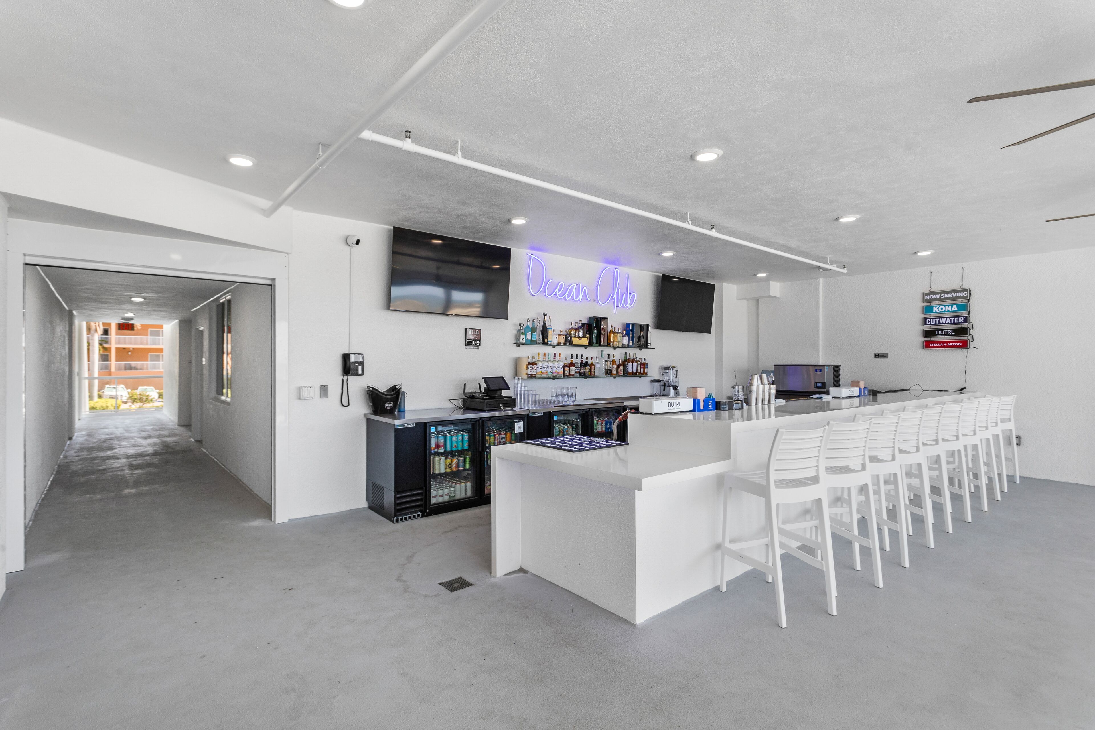 Un área de bar moderna con un letrero de neón, una barra, taburetes y un refrigerador lleno de bebidas.