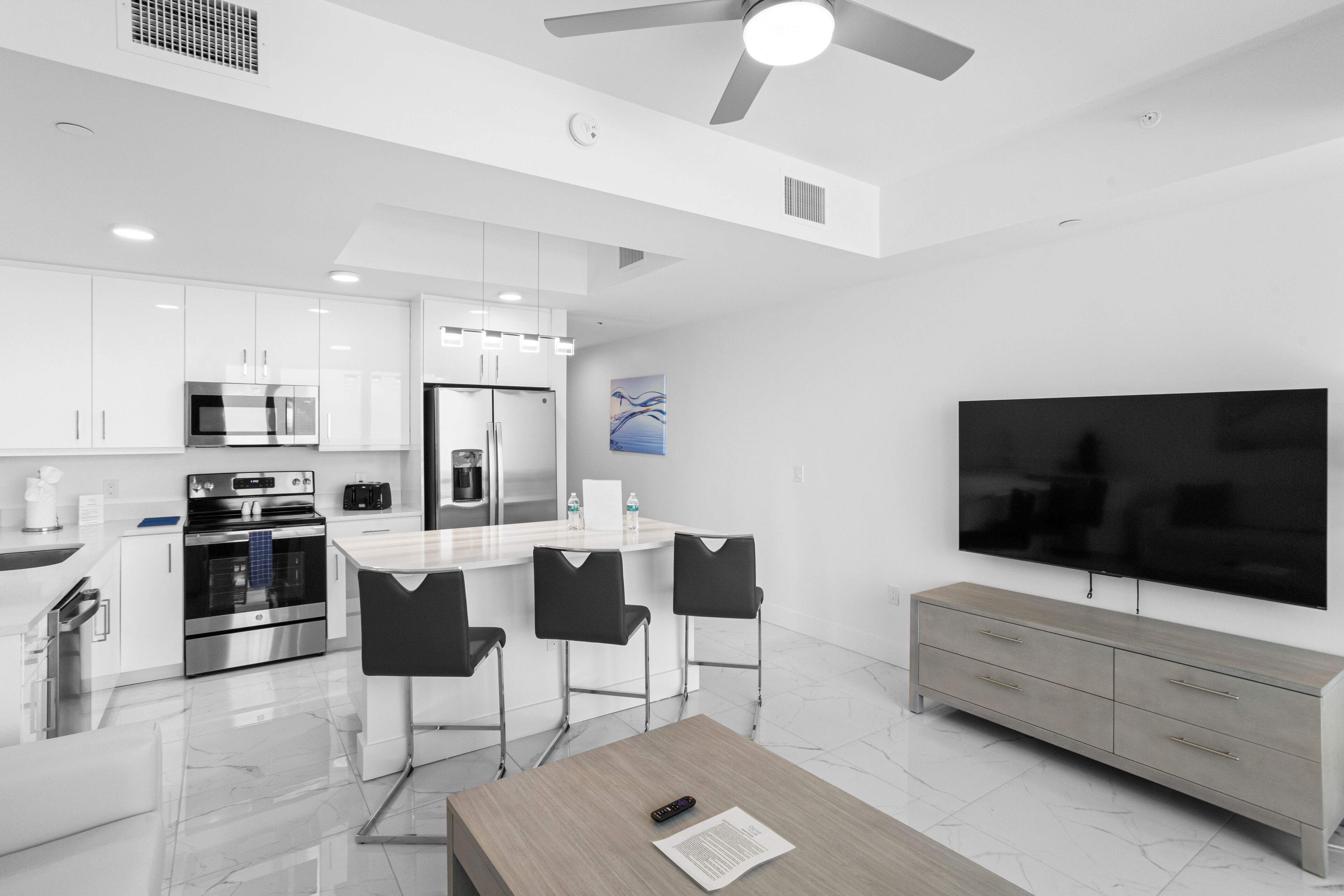 A modern kitchen with white cabinets, a stainless steel refrigerator, and a built-in oven. There is a dining area with a table and chairs, and a large flat-screen TV mounted on the wall.